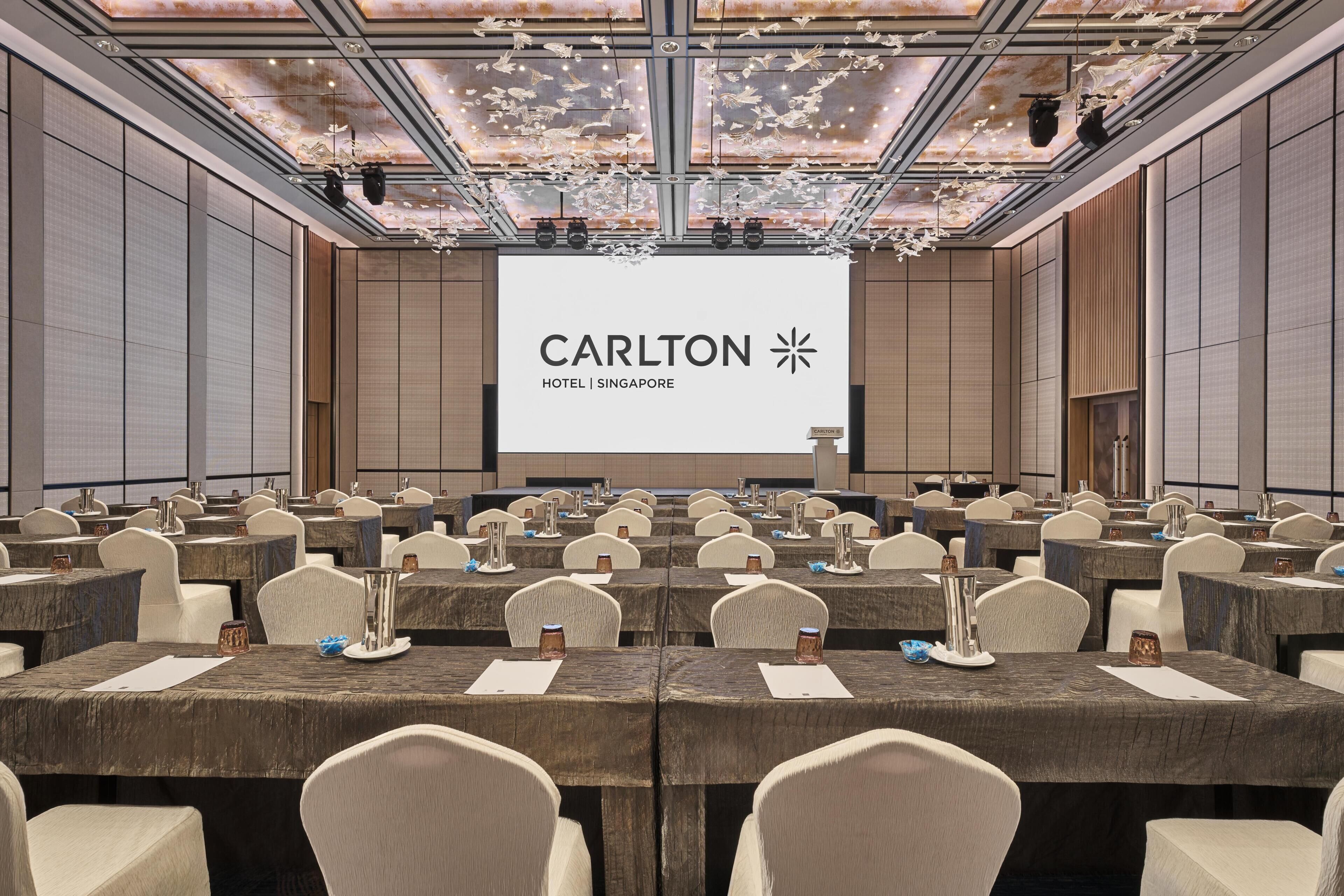 A conference room with multiple tables and chairs, a large screen displaying 'Carlton Hotel Singapore', and a ceiling with a unique light fixture.