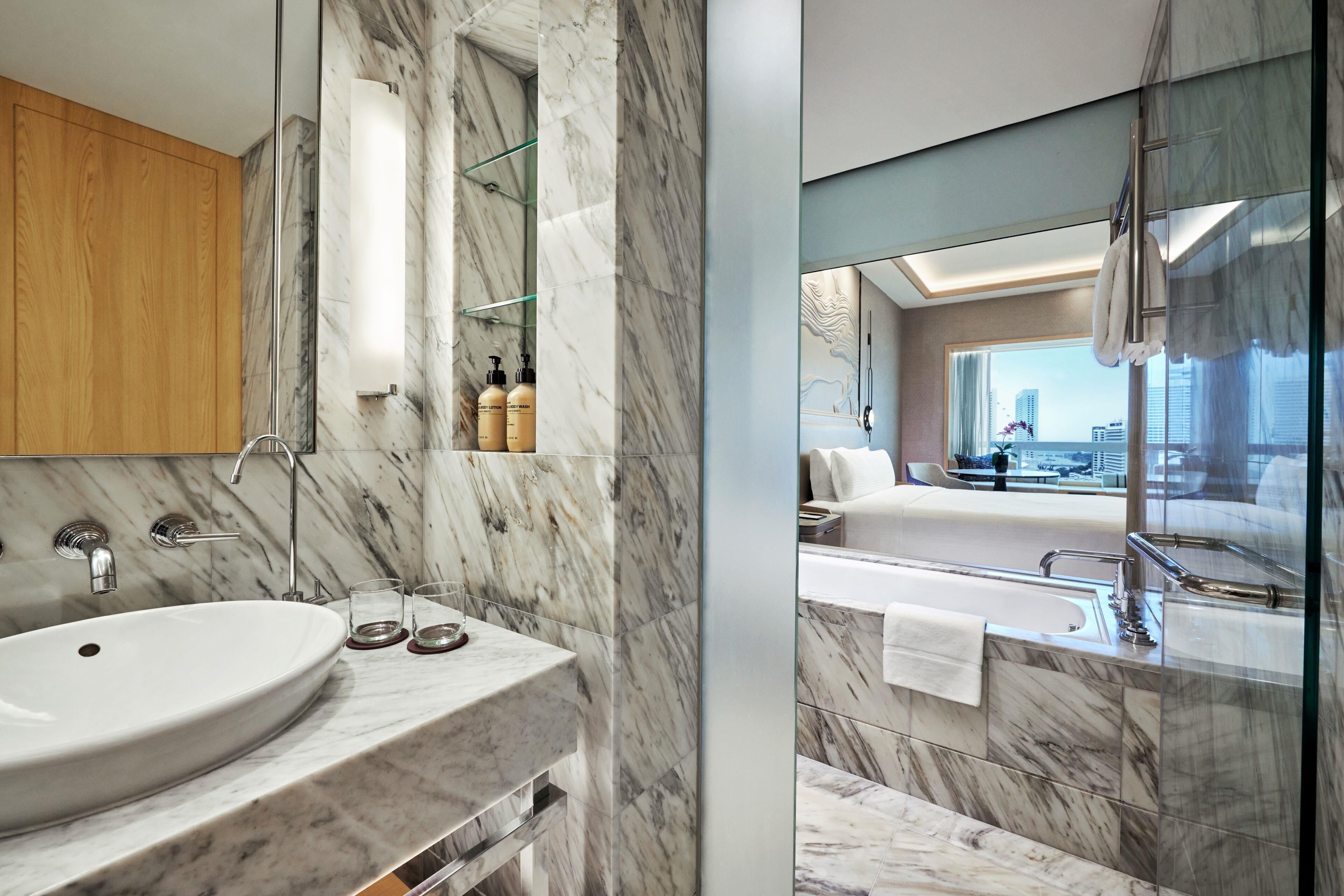 A modern bathroom with a marble sink, a glass shower enclosure, and a view of a cityscape through a large window.