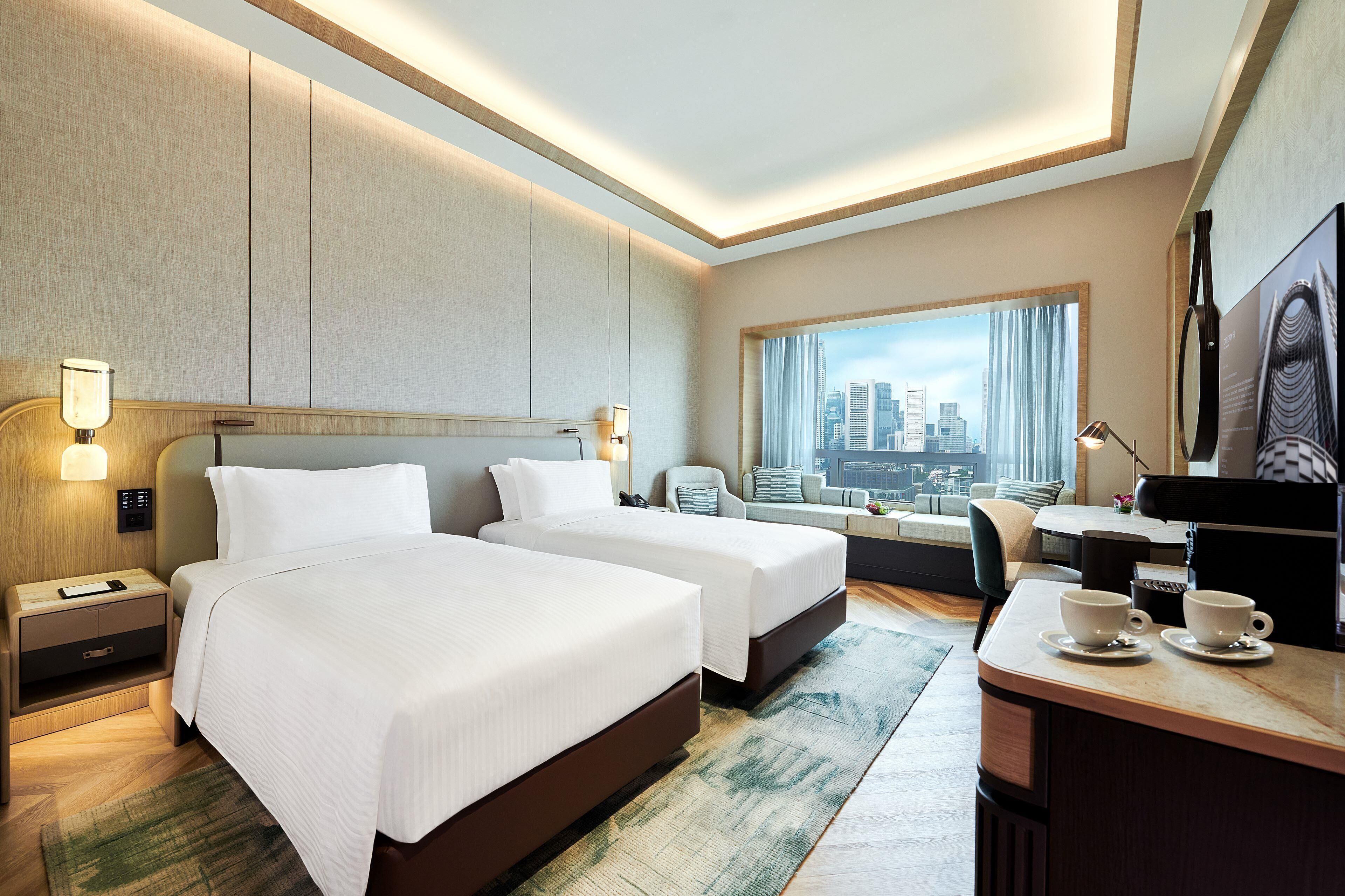 Habitación de hotel con dos camas, un escritorio con silla, televisión y vistas a la ciudad.