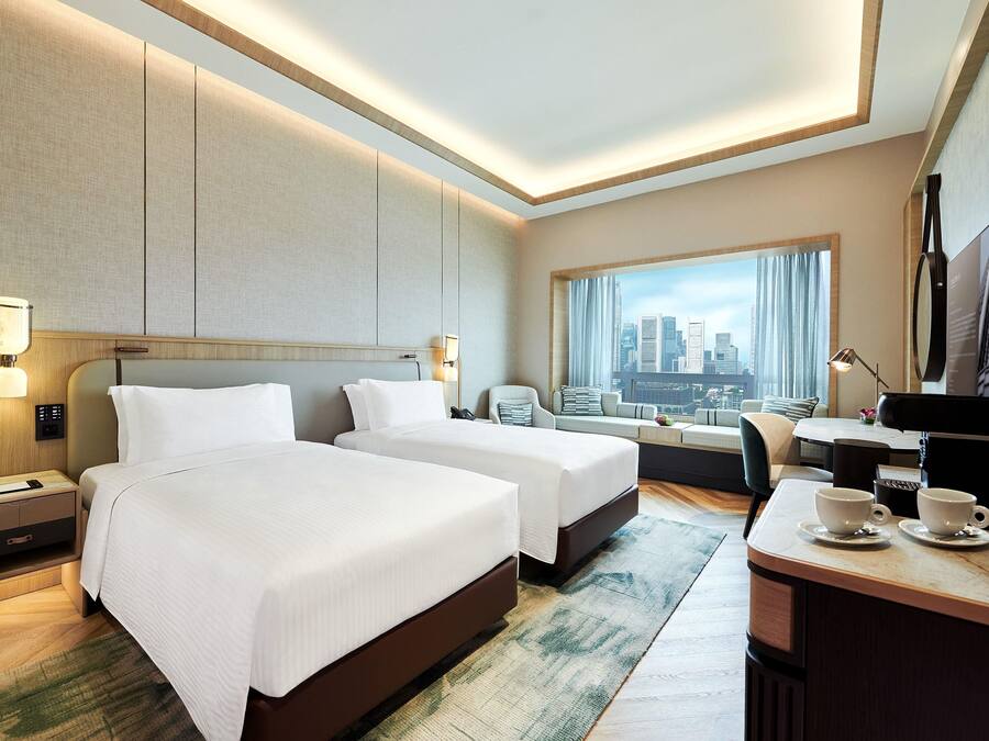 Habitación de hotel con dos camas, un escritorio con silla, televisión y vistas a la ciudad.
