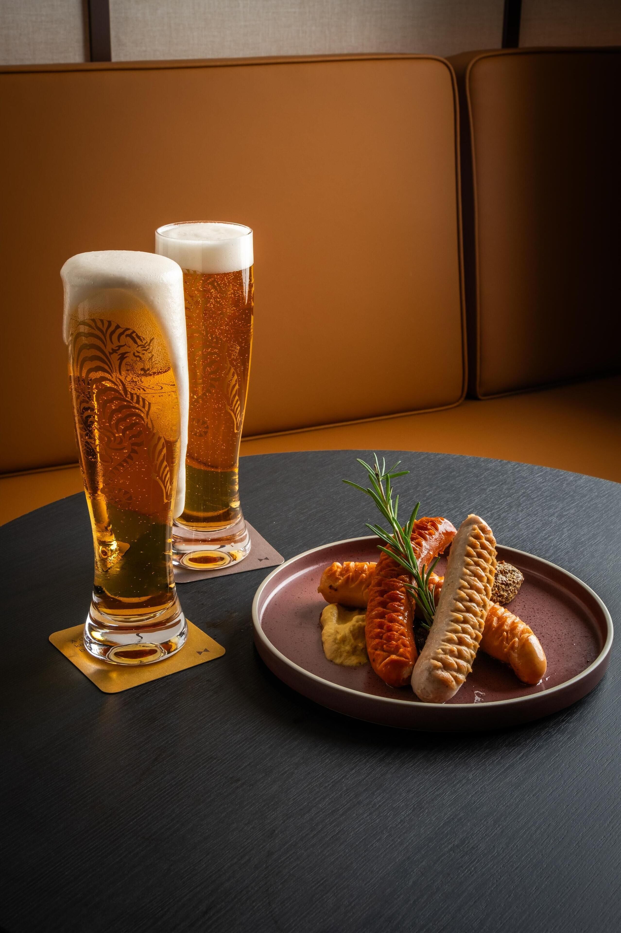 Two tall glasses of beer on a coaster, a plate with pretzels and a sprig of rosemary, and a dark table.