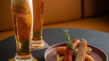 Two tall glasses of beer on a coaster, a plate with pretzels and a sprig of rosemary, and a dark table.