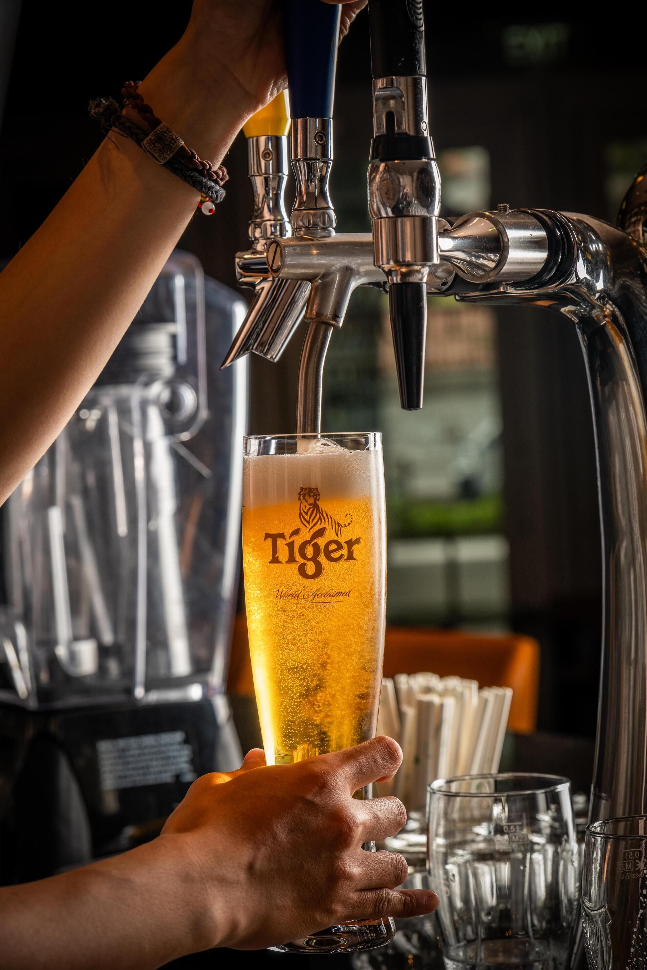 Eine Hand hält ein Glas Bier vor einem Zapfhahn mit der Aufschrift 'Tiger'.