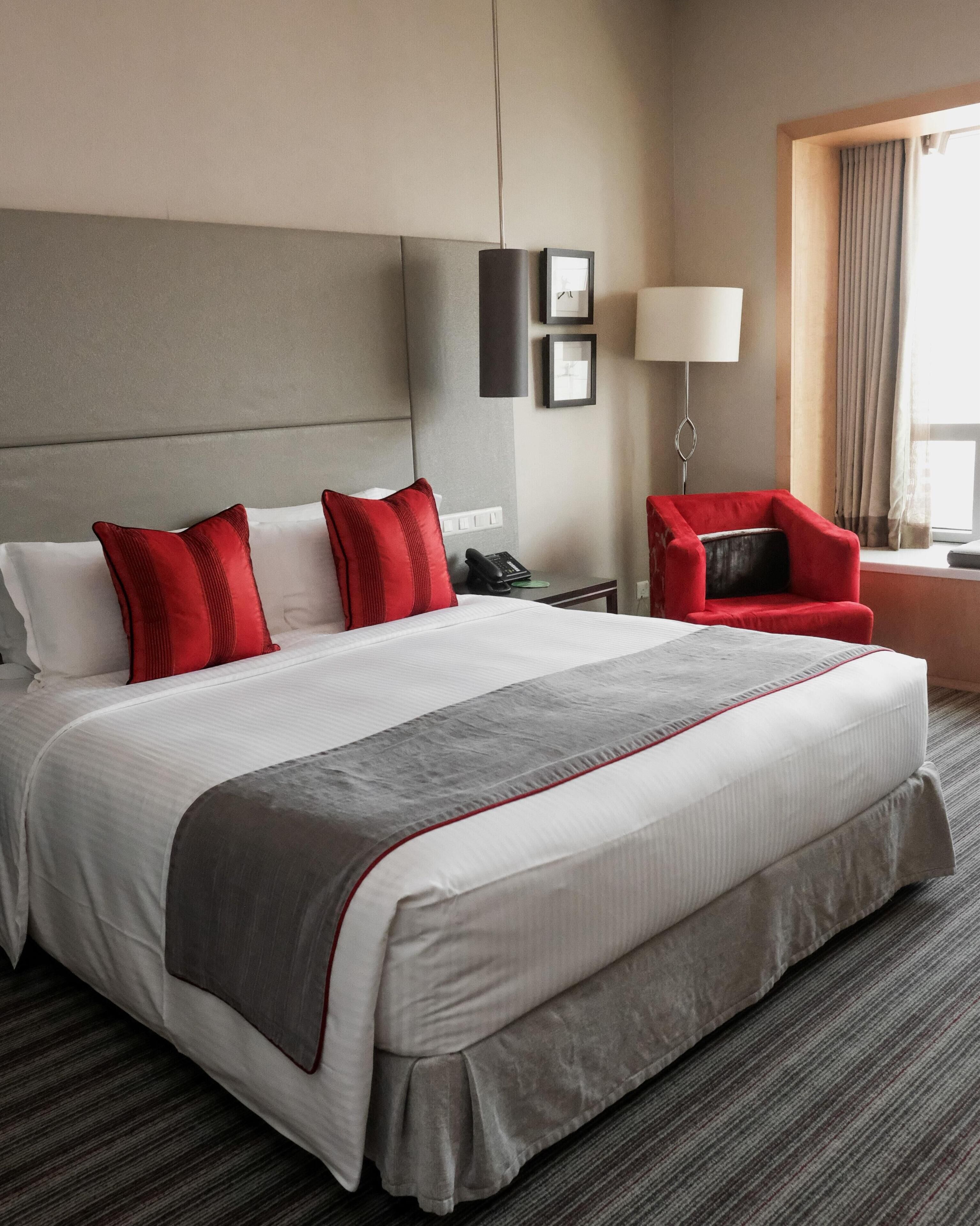 A hotel room with a large bed, a red chair, a desk with a phone, and a lamp.