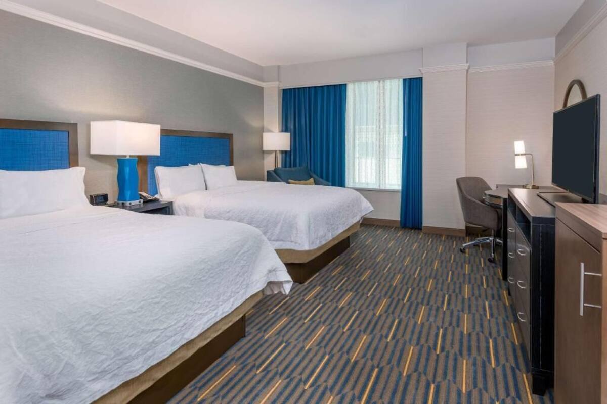 A hotel room with two beds, a desk, a TV, and a window with curtains.