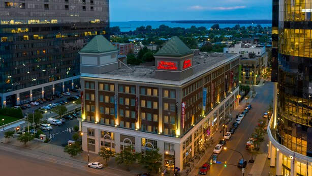 A hotel with a prominent red sign, large windows, and a parking lot with multiple cars. - Hampton Inn & Suites Buffalo Downtown (Buffalo)