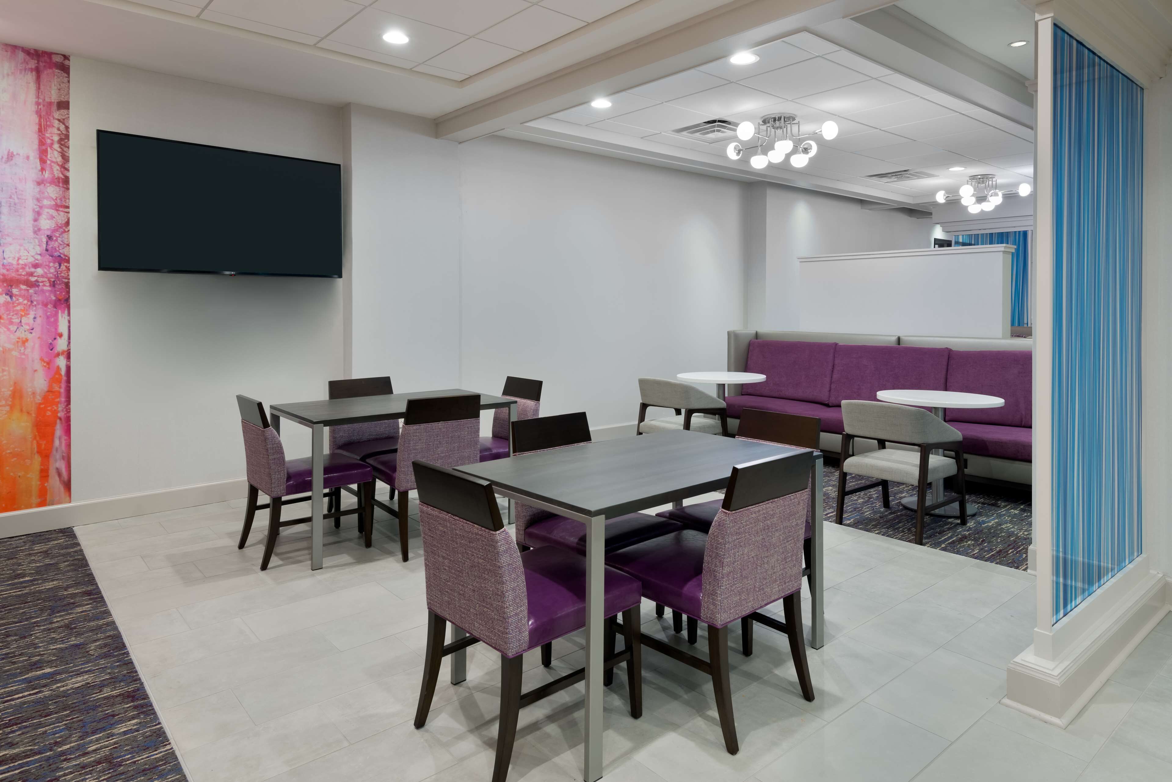 A modern hotel lobby with a flat-screen TV mounted on the wall, a reception desk, purple seating, and a patterned rug.