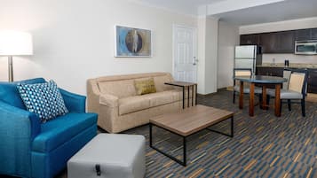 Et hotelværelse med en blå sofa, en beige sofa, et lille bord, en grå skammel, et maleri på væggen, en hvid dør og et tekøkken med mikroovn og køleskab.