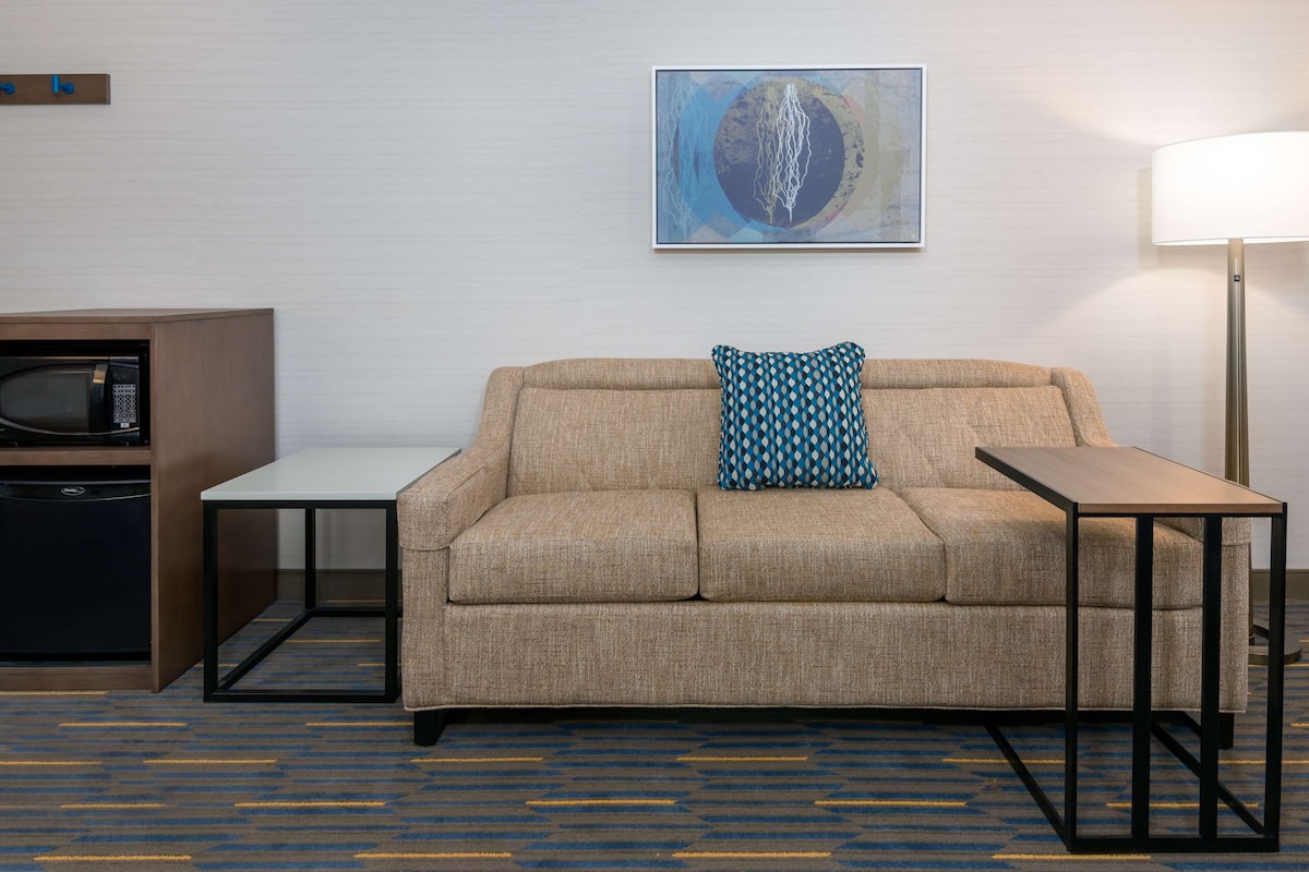 A hotel room with a beige sofa, a patterned pillow, a small table, a microwave, and a lamp.