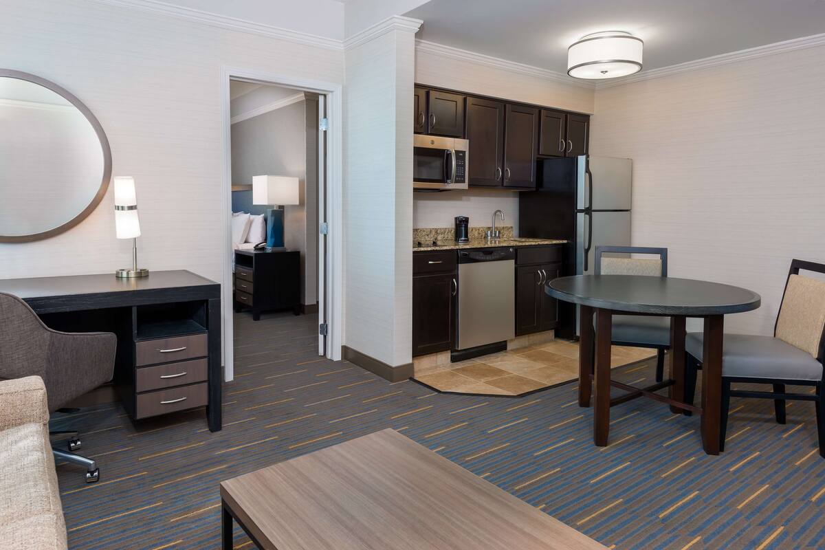 A hotel room with a desk, chair, dining table, and a kitchenette.