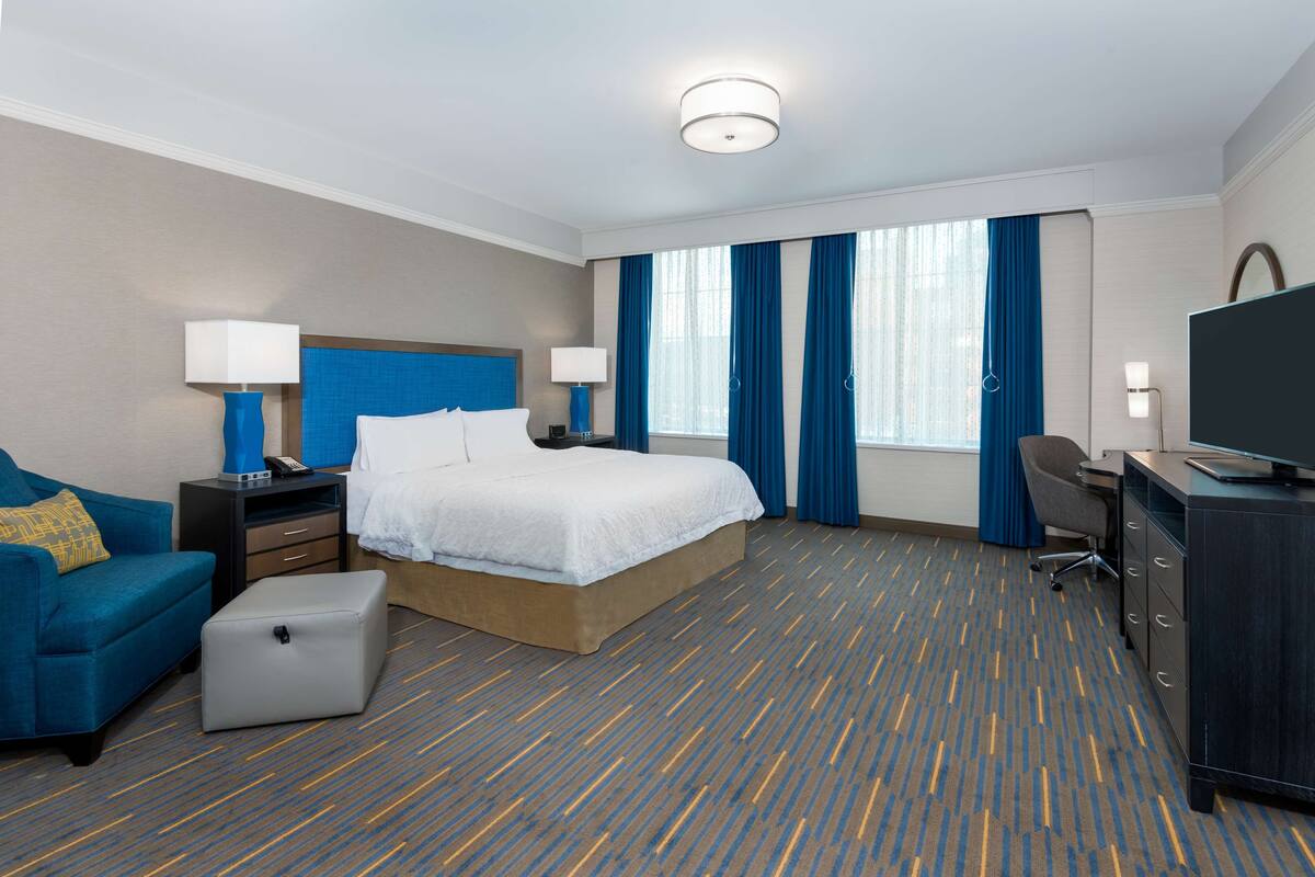 A hotel room with a bed, a desk, a chair, a sofa, a TV, and a window with curtains.