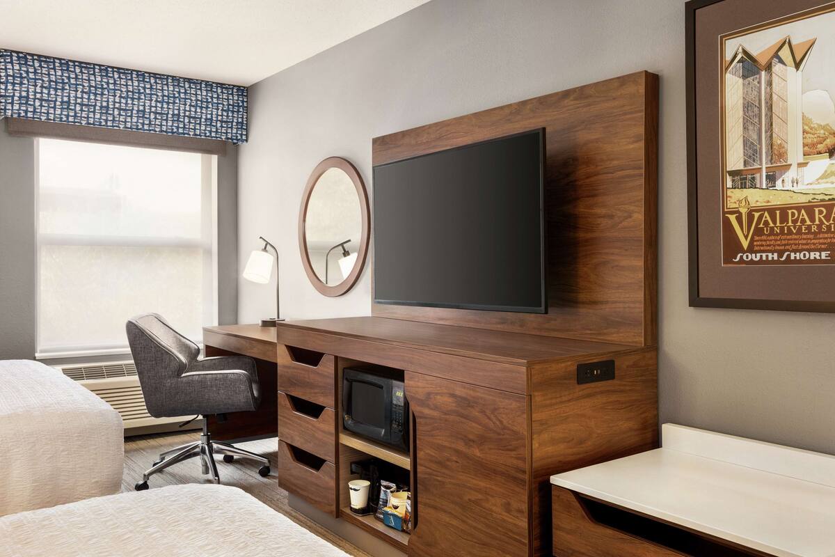 A hotel room with a wooden desk, a flat-screen TV, a chair, a microwave, and a large window.