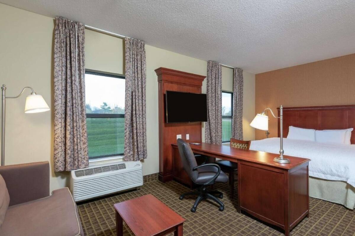 A hotel room with a bed, desk, chair, lamp, TV, and a window with curtains.