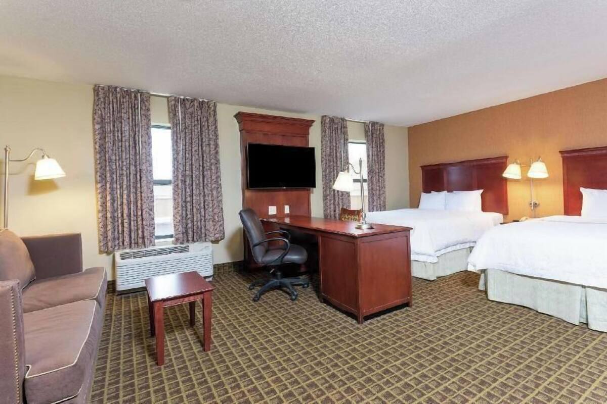 A hotel room with two beds, a desk, a chair, a TV, and a lamp.