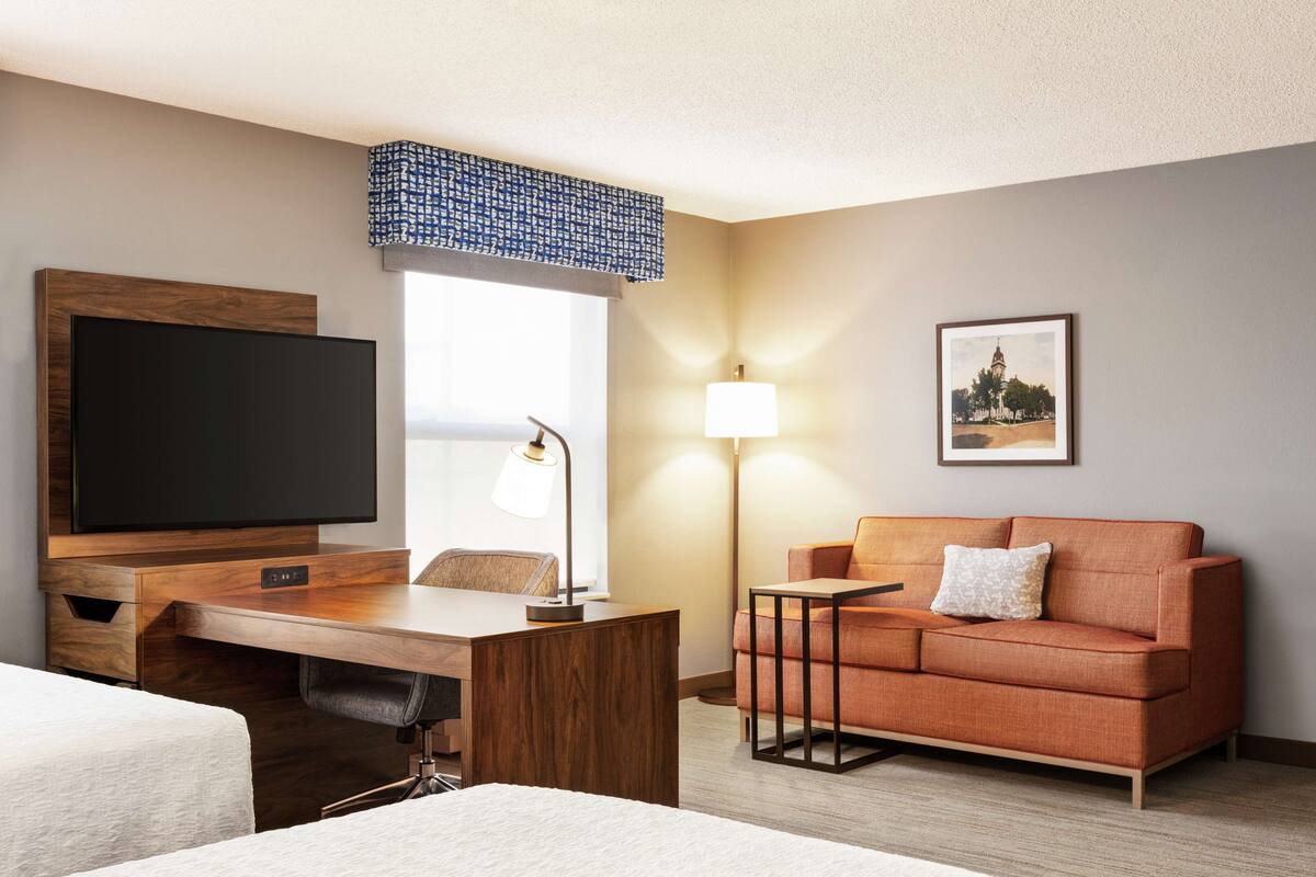 A hotel room with a wooden desk, a television, a sofa, a lamp, and a framed picture on the wall.