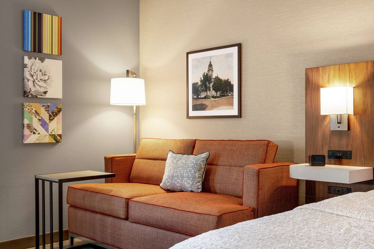 A hotel room with a sofa, a bedside table, a lamp, a framed picture, and a nightstand with a lamp.