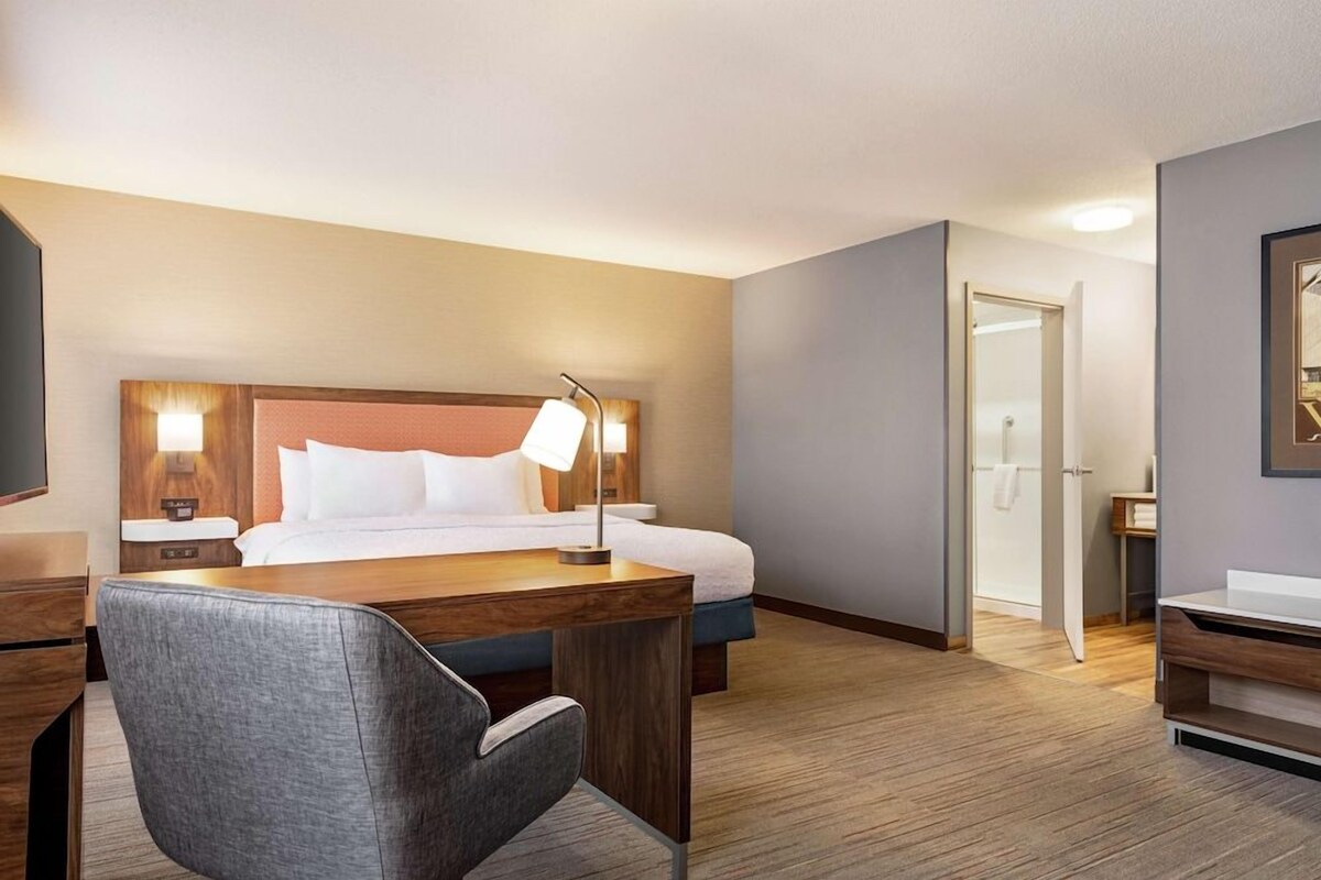 Habitación de hotel con cama, escritorio con silla, mesita de noche con lámpara y baño visible a través de una puerta abierta.