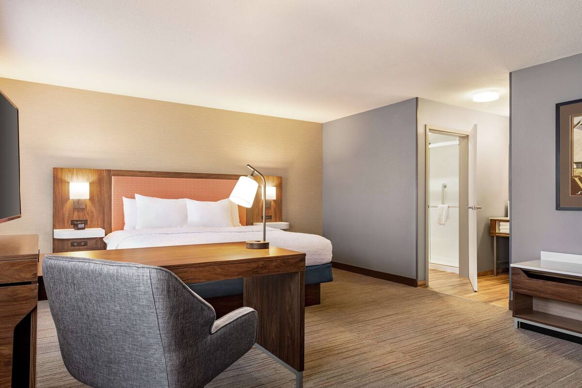 A hotel room with a wooden desk, a chair, a bed with white linens, a nightstand with a lamp, and a door leading to a bathroom.
