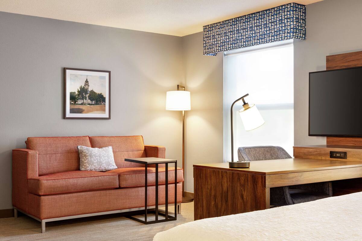 A hotel room with a sofa, a desk, a lamp, a TV, and a window with a patterned curtain.