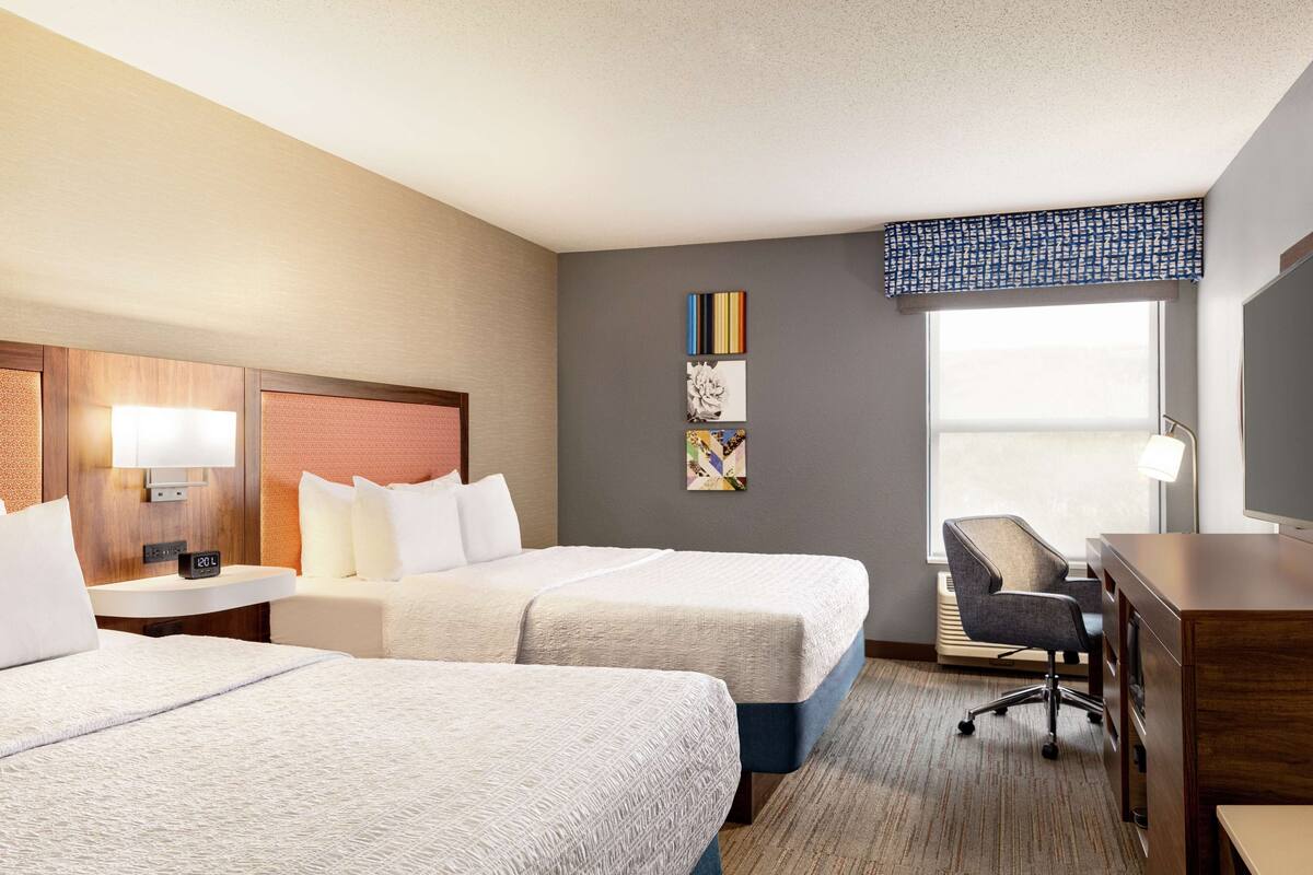 A hotel room with two beds, a desk, a chair, a lamp, and artwork on the wall.