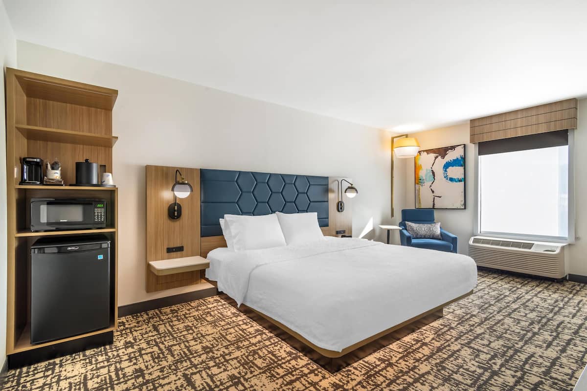 A hotel room with a bed, a microwave, a refrigerator, a wooden shelf, a headboard, a lamp, a chair, a window, and a wall art piece.