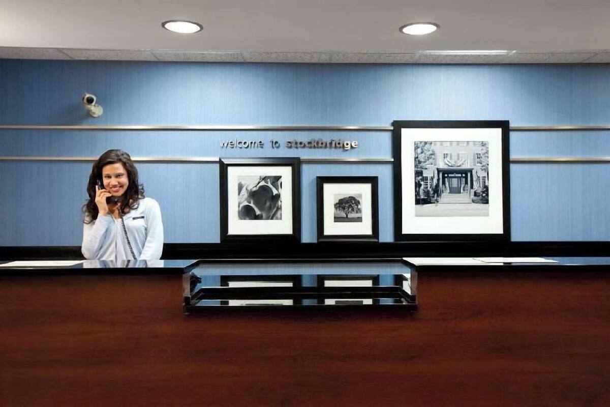 A reception desk with a 'Welcome to Stockbridge' sign, framed black and white photographs on the wall, and a security camera above.