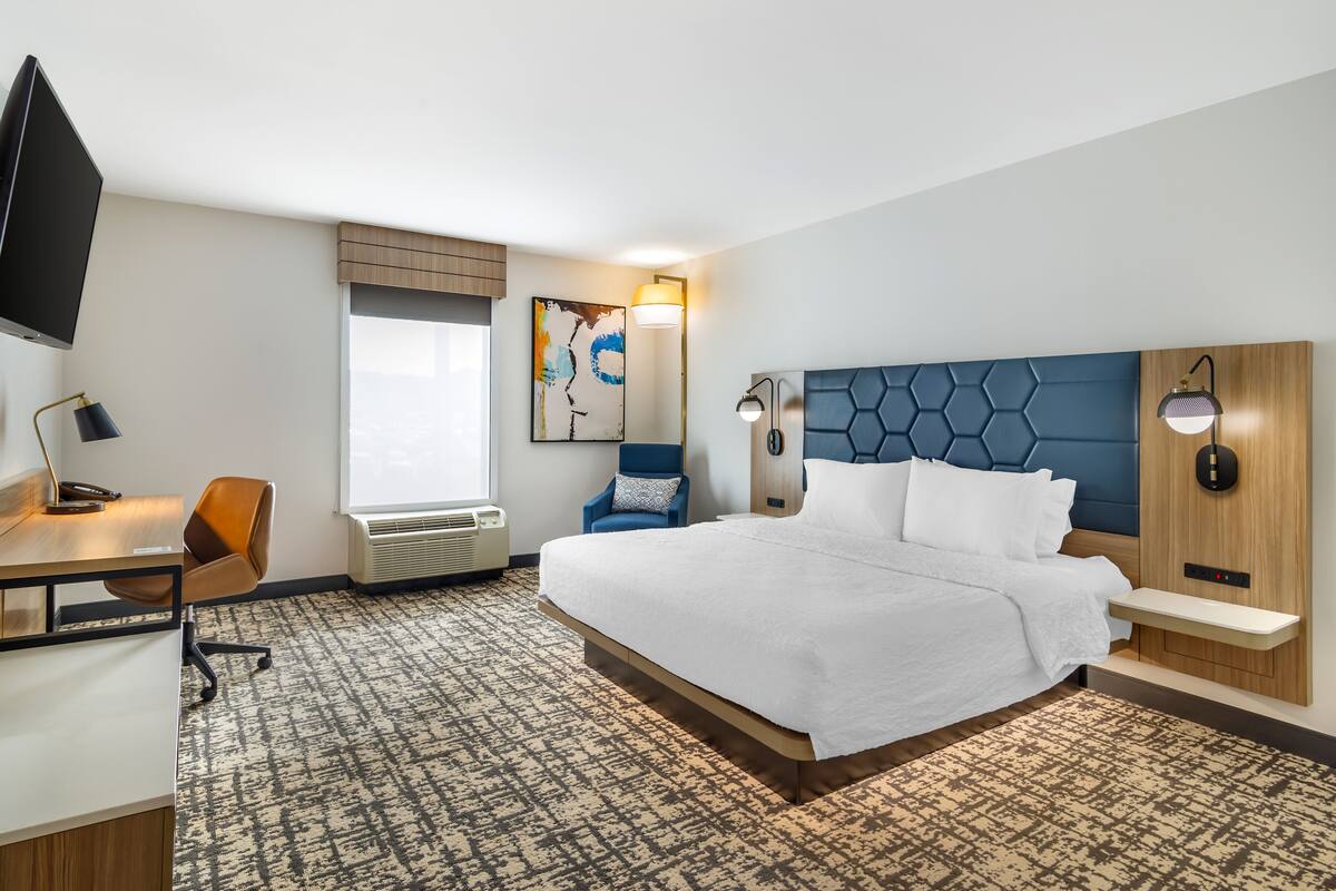 A hotel room with a large bed, a desk with a chair, a TV, a window, and a wall-mounted artwork.