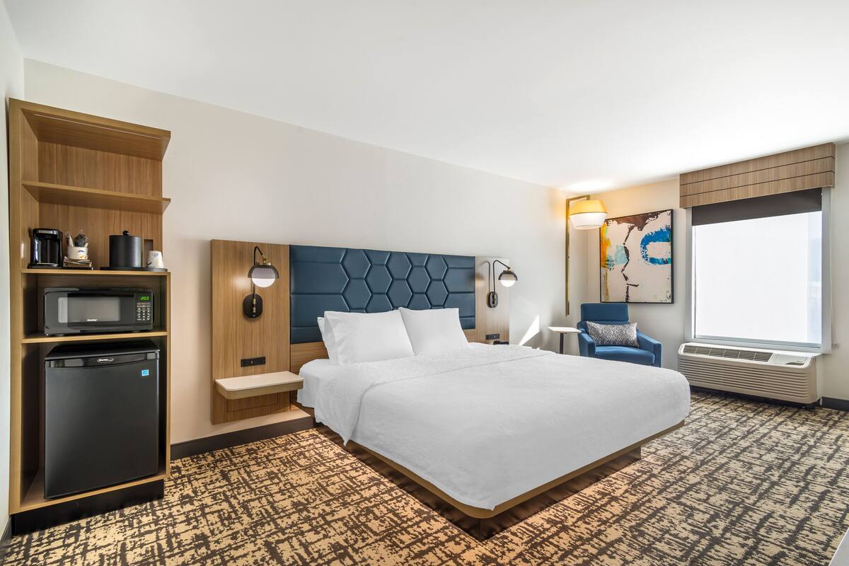 A hotel room with a large bed, a built-in microwave, a refrigerator, a wooden shelf, a headboard with a geometric pattern, a bedside table with a lamp, a chair, and a large window.