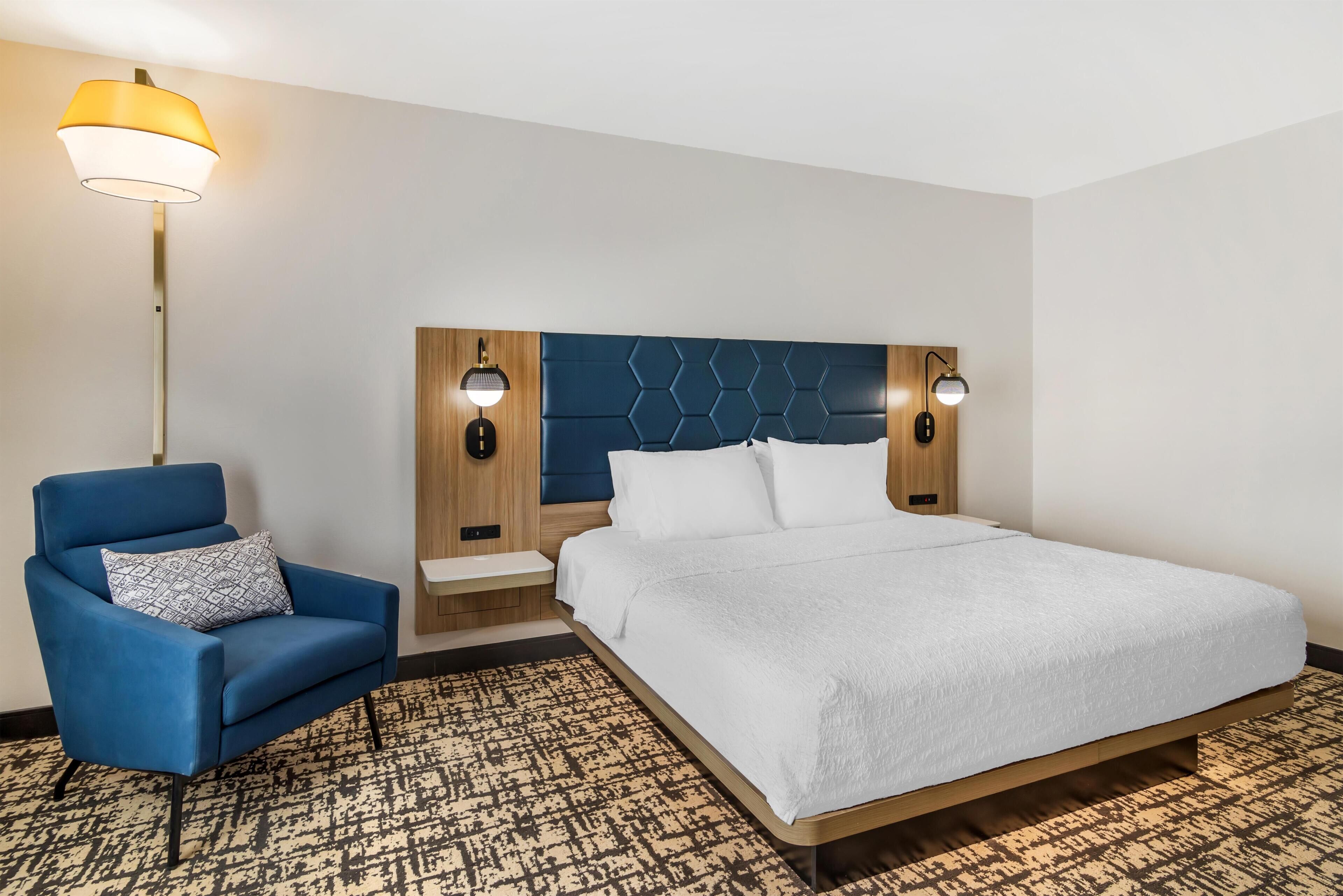 Ett hotellrum med en säng, en blå fåtölj, en mönstrad matta och en modern sänggavel med väggmonterade lampor.