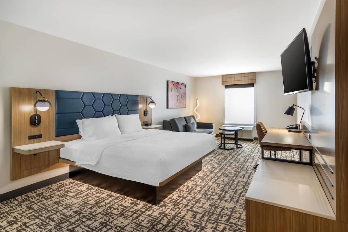 A modern hotel room with a large bed, a desk, a TV, and a seating area.