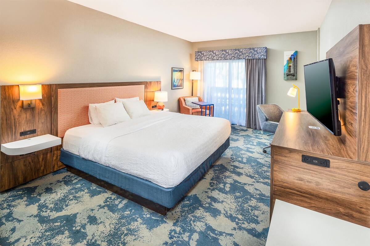 Quarto de hotel com uma cama grande, mesas de cabeceira, escrivaninha com cadeira, televisão e janela com cortinas.