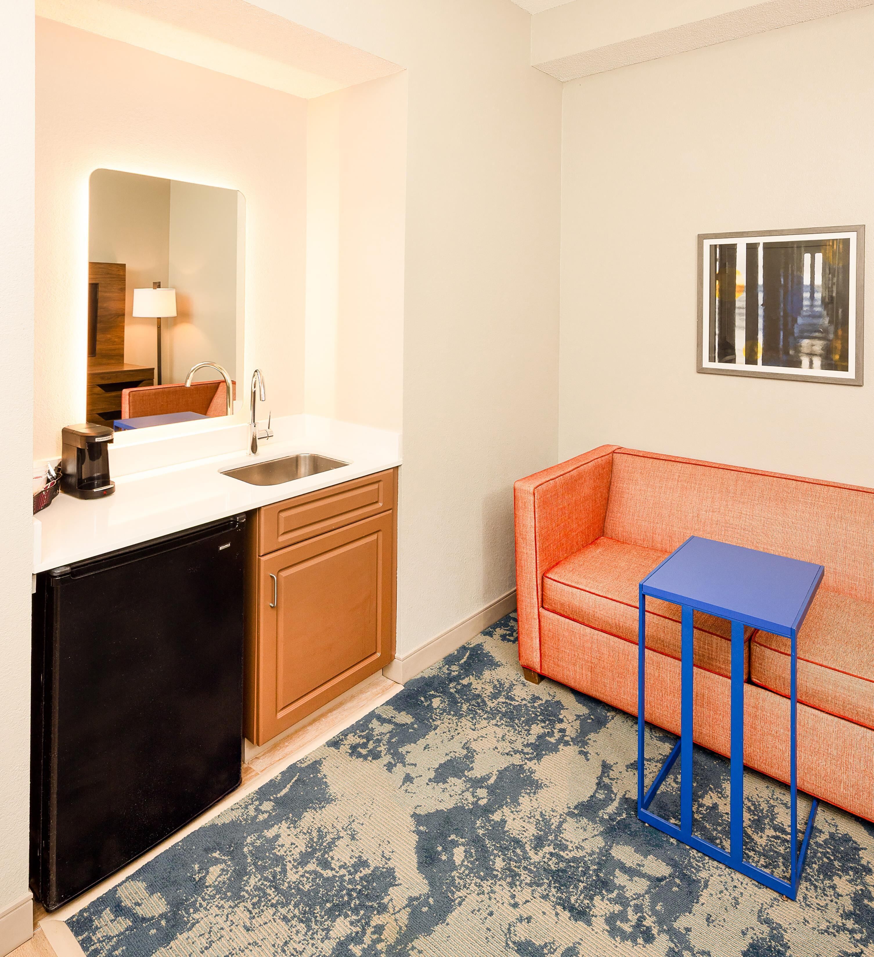 Een compacte kitchenette met een spoelbak, een koffiezetapparaat en een kleine koelkast. Er staat een blauwe tafel naast een oranje bank.