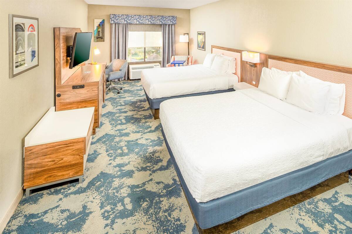 Quarto de hotel com duas camas, uma escrivaninha, uma cadeira, uma televisão e uma janela com cortinas.