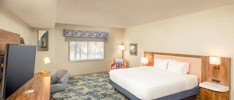 A hotel room with a large bed, a desk with a computer, a chair, and a window with a view of trees.