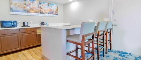 A kitchen with a white countertop, wooden cabinets, and a microwave. There are three wooden chairs with light-colored cushions. A painting of a coastal scene hangs above the counter.