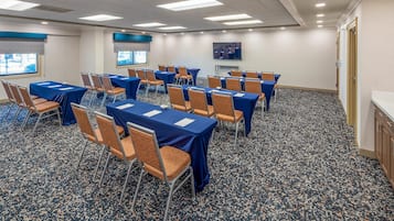 A conference room with tables and chairs, carpeted floor, and a view of the outdoors through windows.