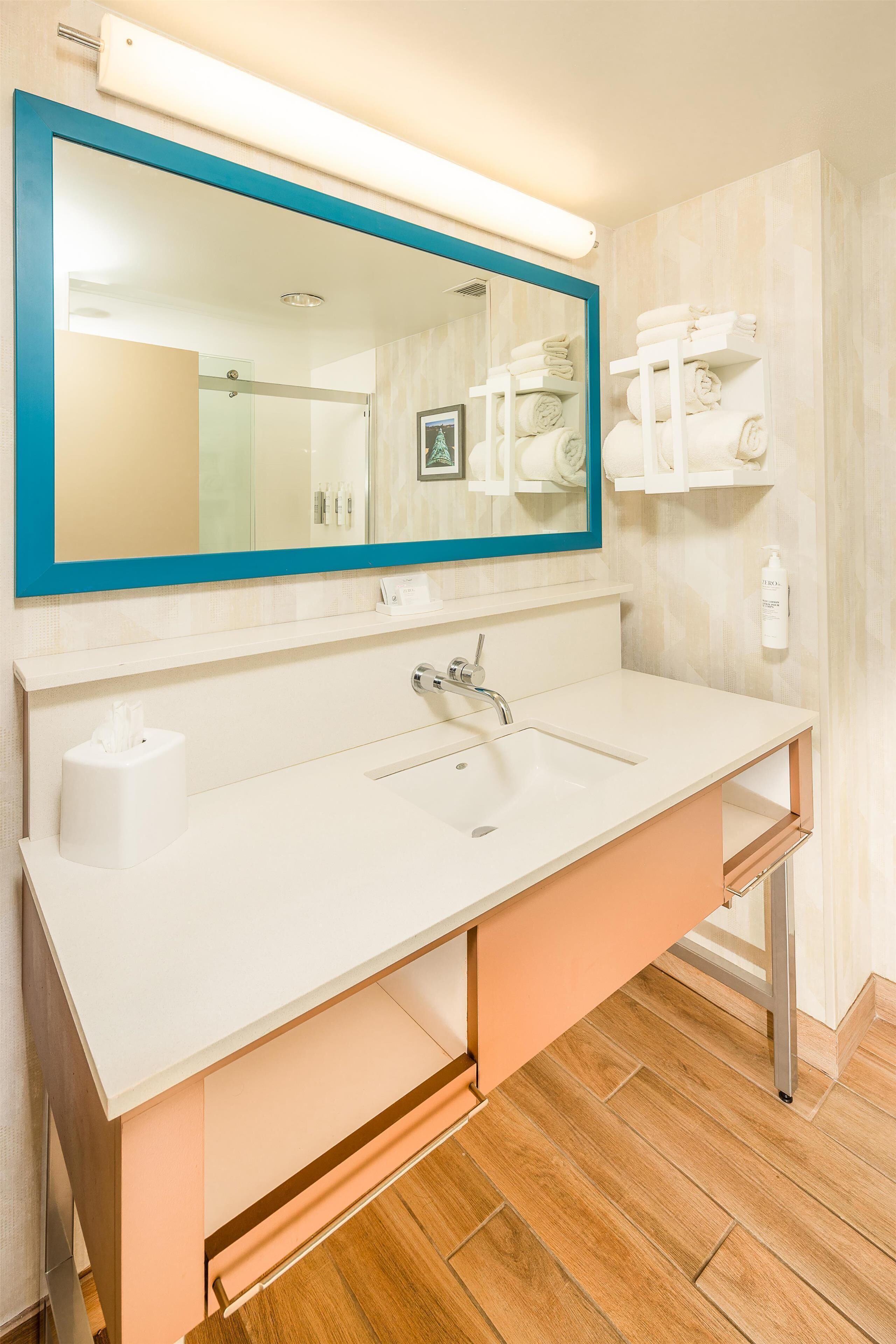 A modern bathroom with a white sink, a blue-framed mirror, and neatly arranged towels on shelves.