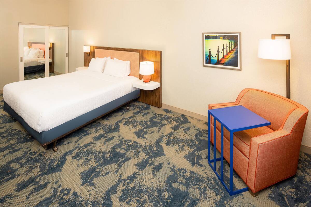 Quarto de hotel com uma cama grande, uma mesinha de cabeceira com abajur, uma pequena mesa, uma poltrona laranja e um quadro na parede.