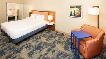 A hotel room with a large bed, a nightstand with a lamp, a small table, an orange armchair, and a painting on the wall.