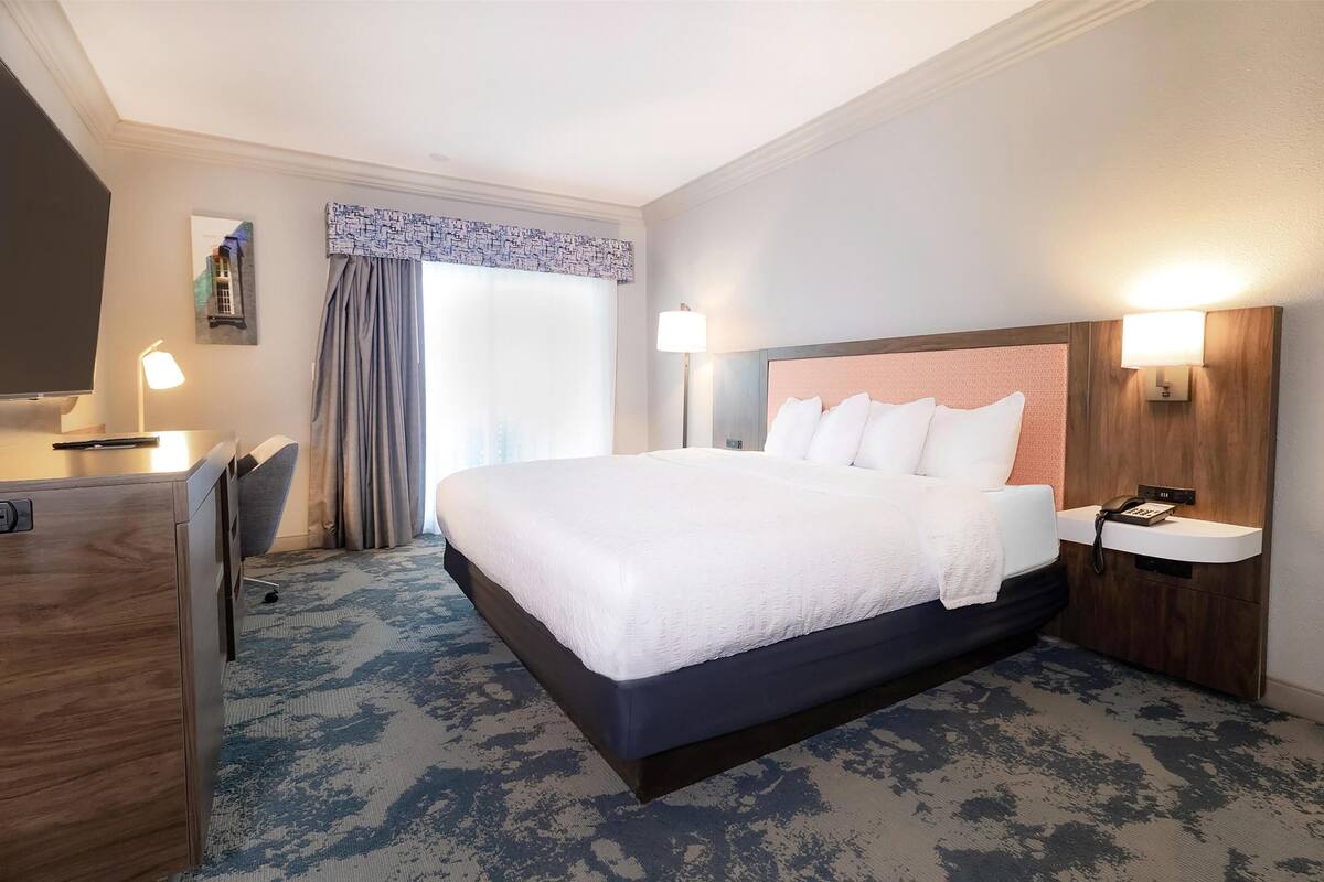 Quarto de hotel com uma cama grande, uma escrivaninha e uma cadeira. Há uma janela com cortinas e uma luminária na parede.