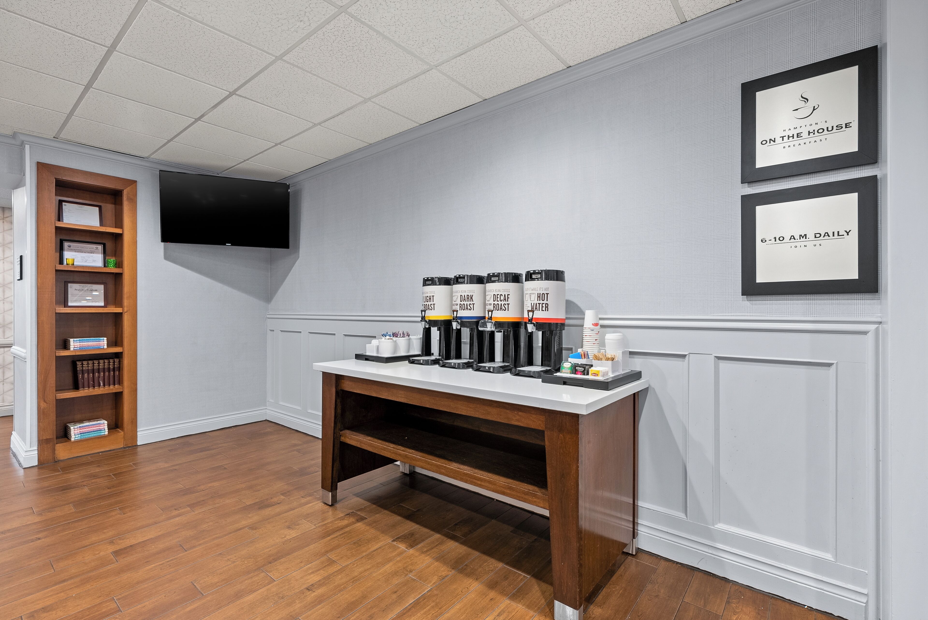 A hotel room with a wooden coffee station, a bookshelf, and a flat-screen TV mounted on the wall.