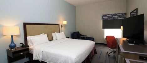 A hotel room with a large bed, a desk with a television, a chair, a lamp, and a nightstand.