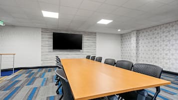 Un salón de conferencias con una larga mesa, sillas negras, un televisor de pantalla plana, paredes con estampado y piso alfombrado.
