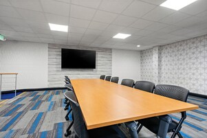 Un salón de conferencias con una larga mesa, sillas negras, un televisor de pantalla plana, paredes con estampado y piso alfombrado.