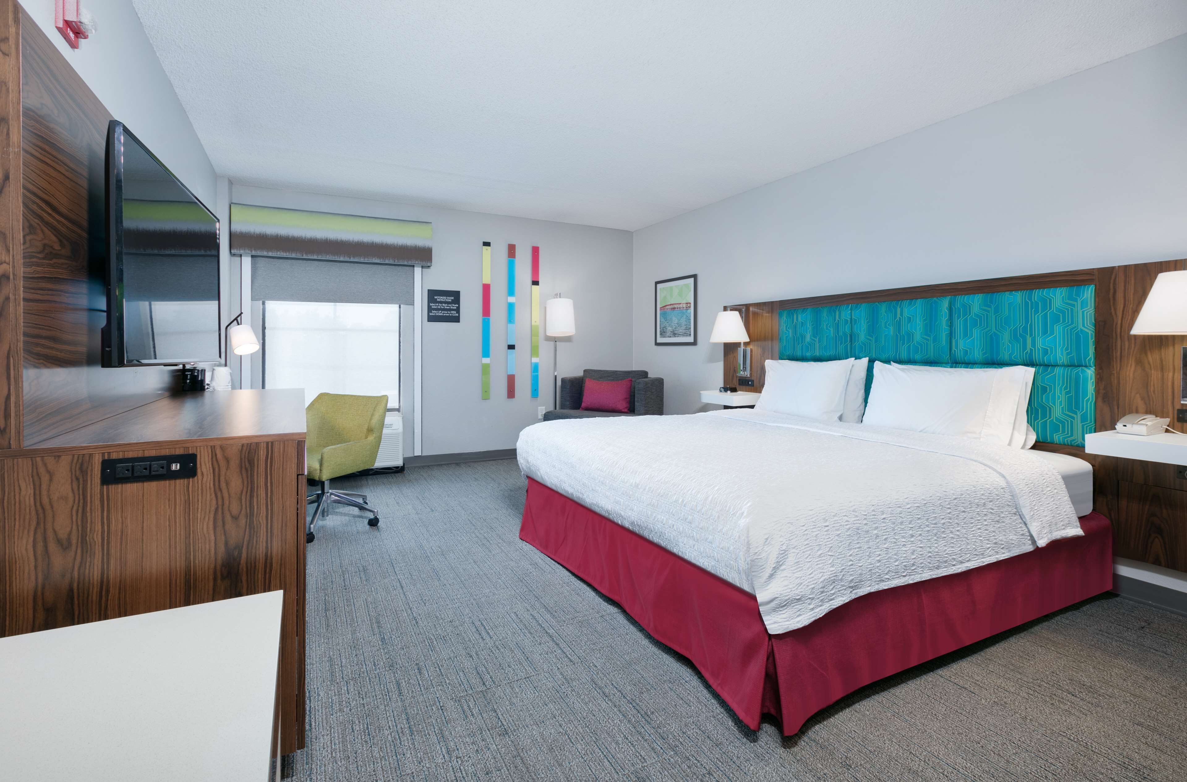 Quarto de hotel com uma cama grande, uma escrivaninha com televisão, uma cadeira e uma luminária de mesa.