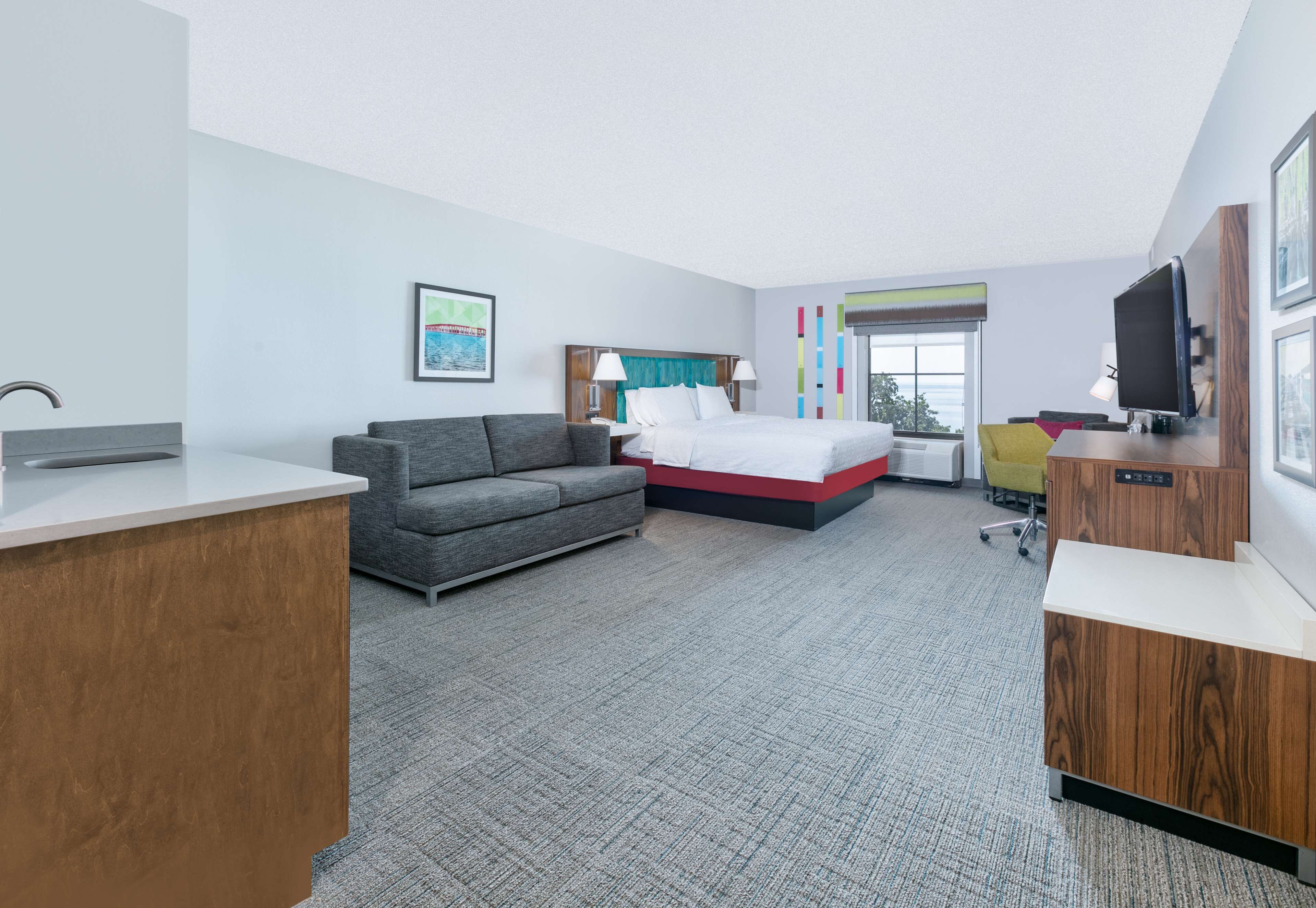 Une chambre d’hôtel moderne avec un lit, un canapé gris, un bureau et un téléviseur.