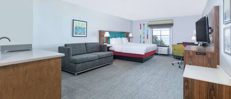 A modern hotel room with a bed, grey sofa, desk, and a TV.
