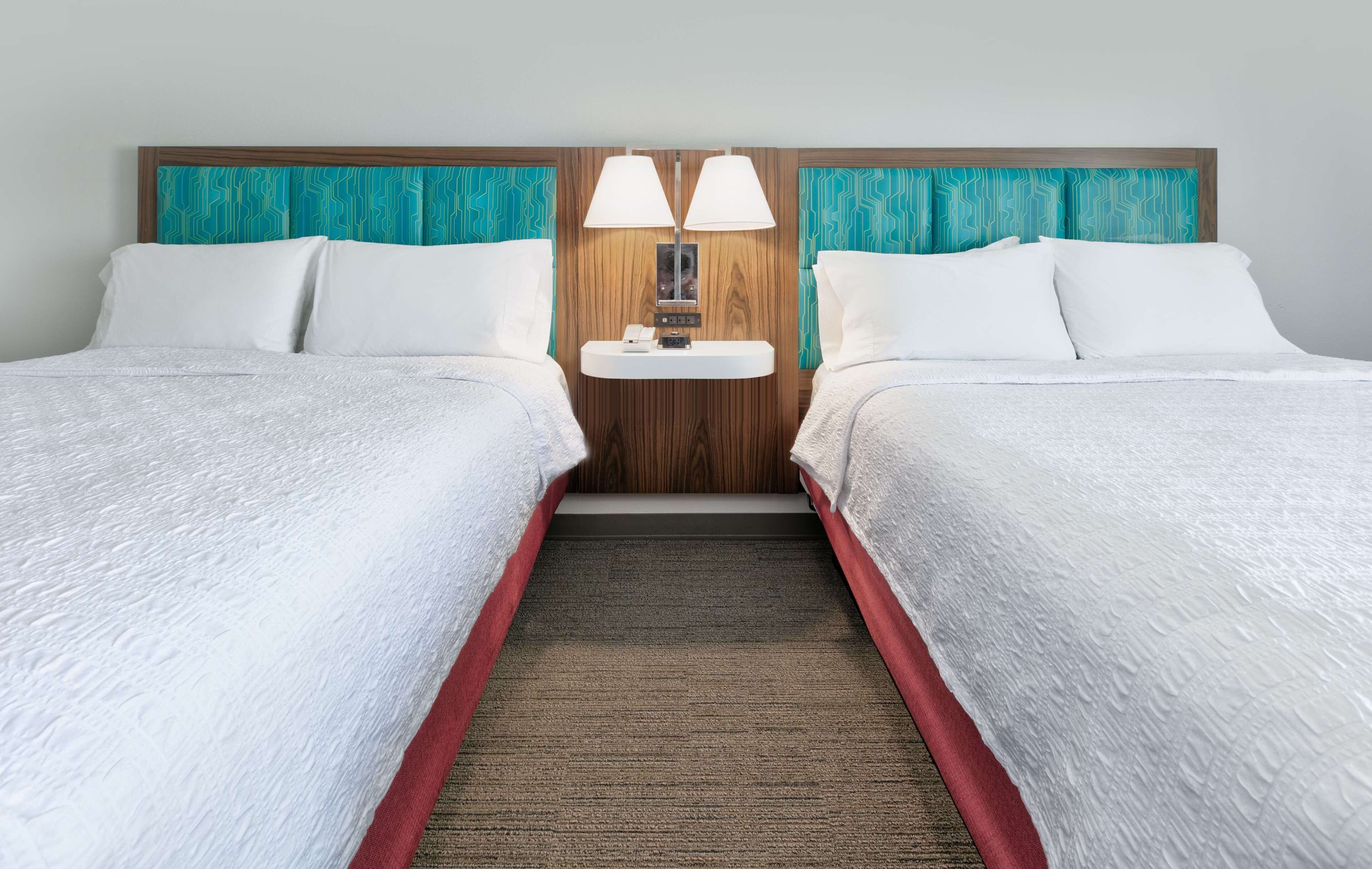 A hotel room with two beds, a wooden headboard, a small table with lamps, and a carpeted floor.