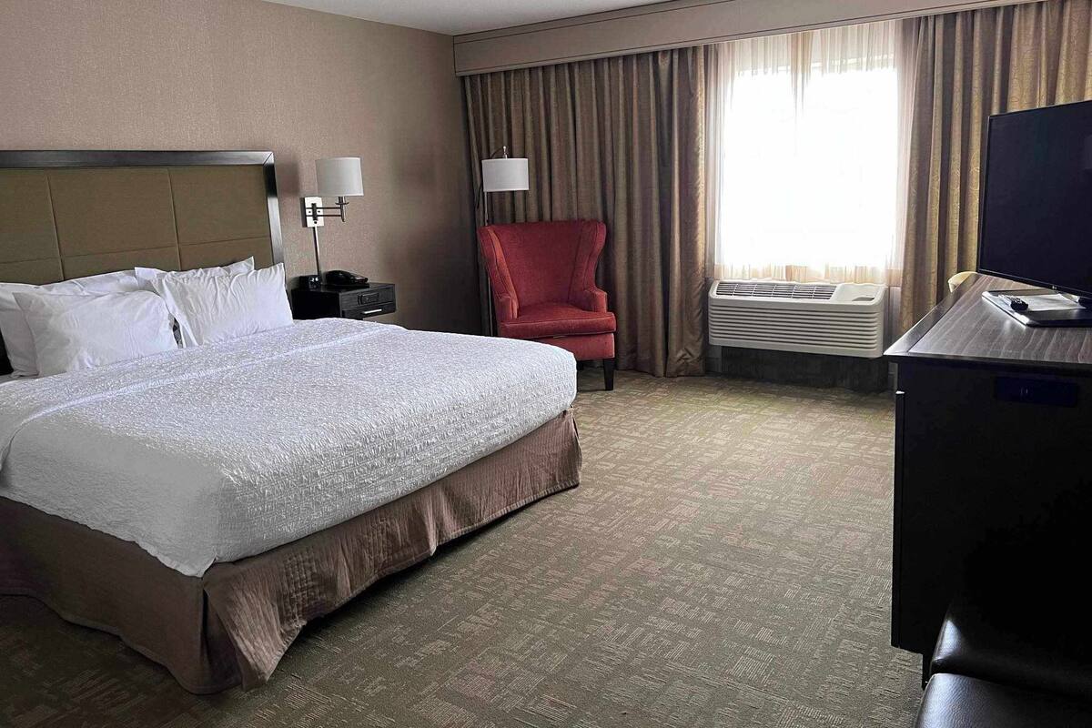 A hotel room with a large bed, a red armchair, a desk with a computer, and a TV.