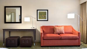 A hotel room with a red sofa, a desk with a lamp, a framed picture on the wall, and a mirror.